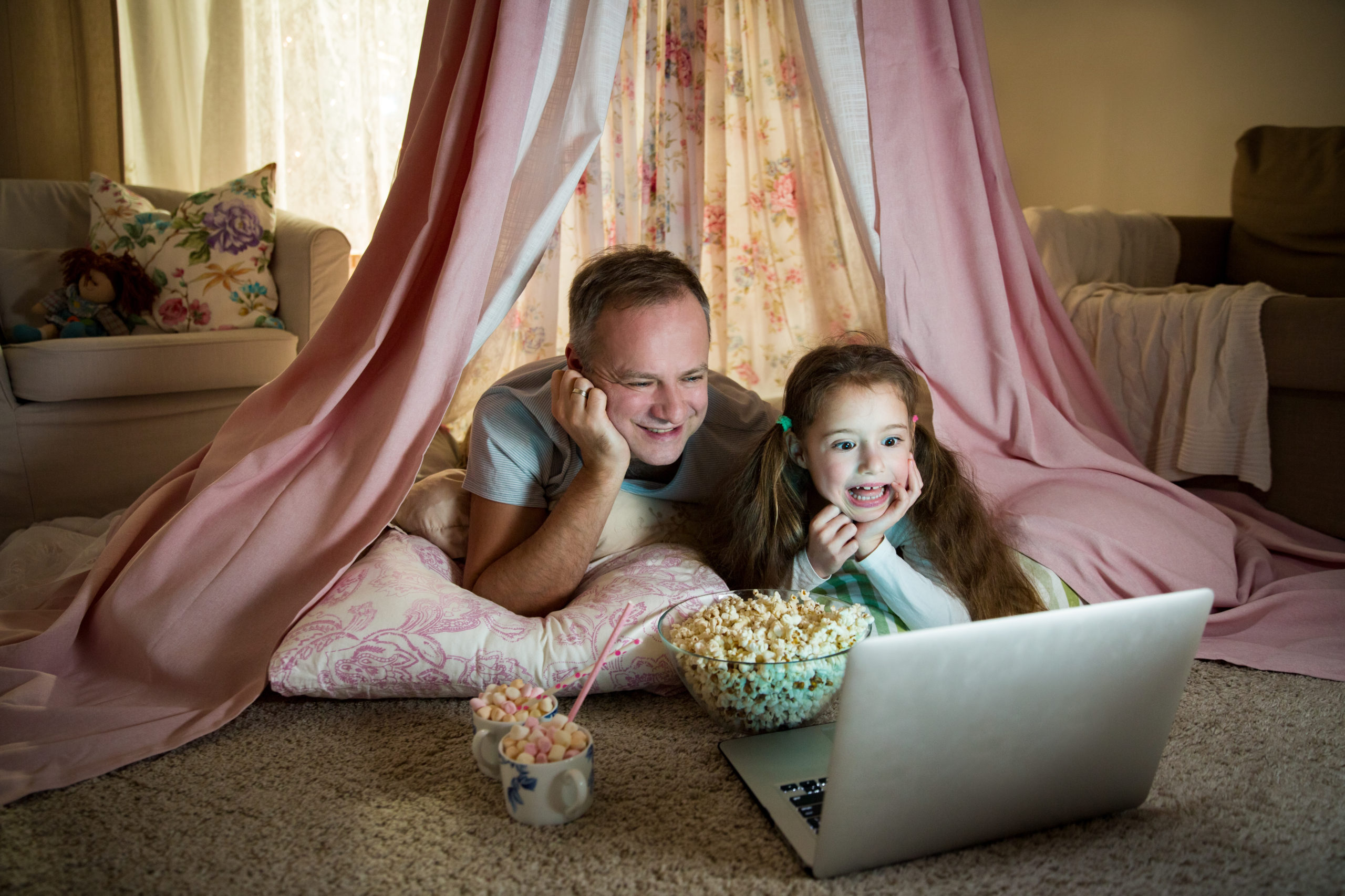 Family quality time. Father and daughter lie in homemade pink tent with flowers, watch cartoons on laptop, eating popcorn, laugh. Cozy stylish room. Family bonds concept